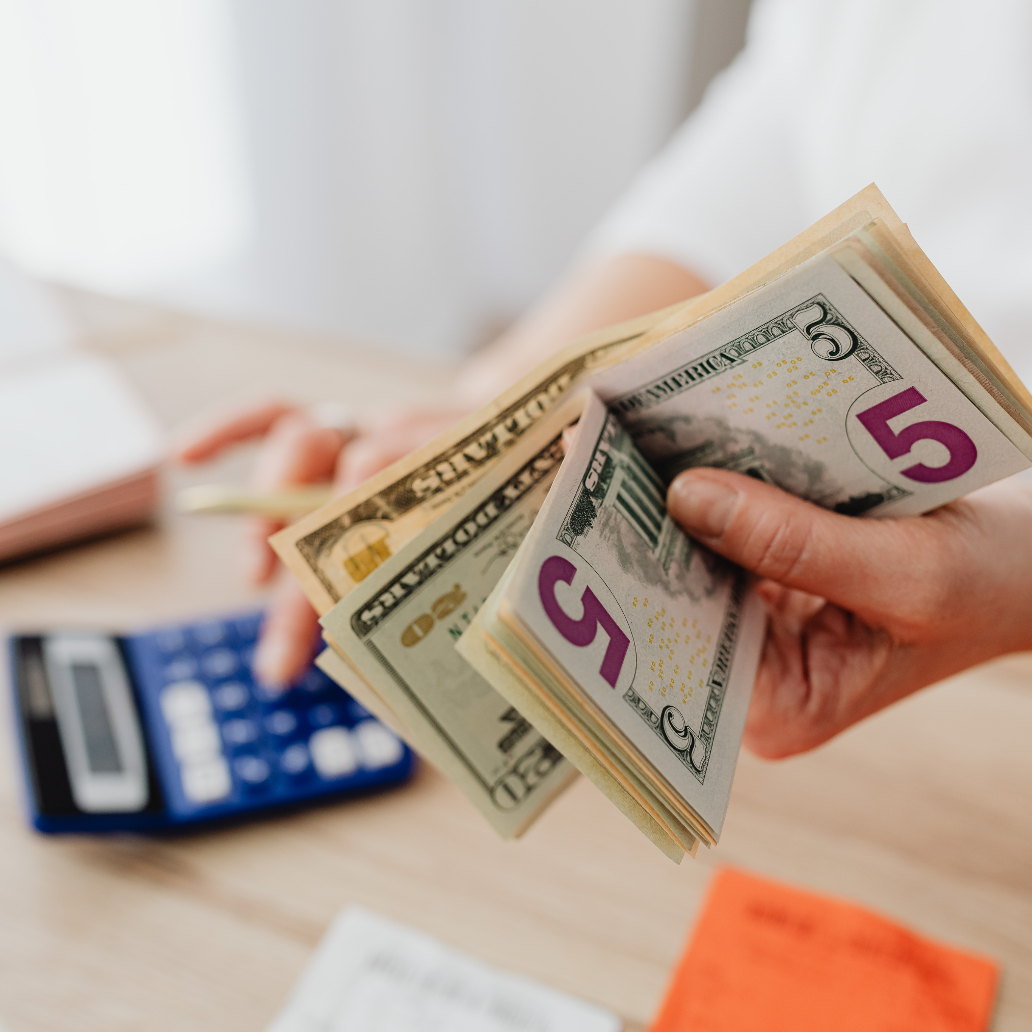 budget - budgetly.money Hands holding a fan of US dollar bills, fives and twenties visible, with a blurred calculator in the background (money exchange or cash handling scene)